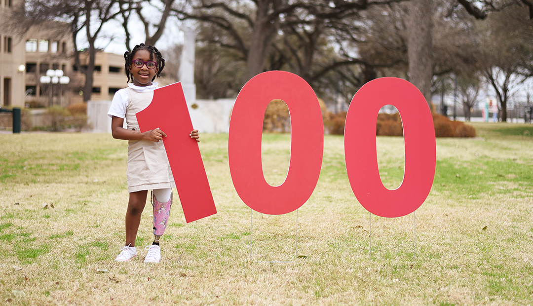 100. A little girl with a prosthetic leg holds up the number 100