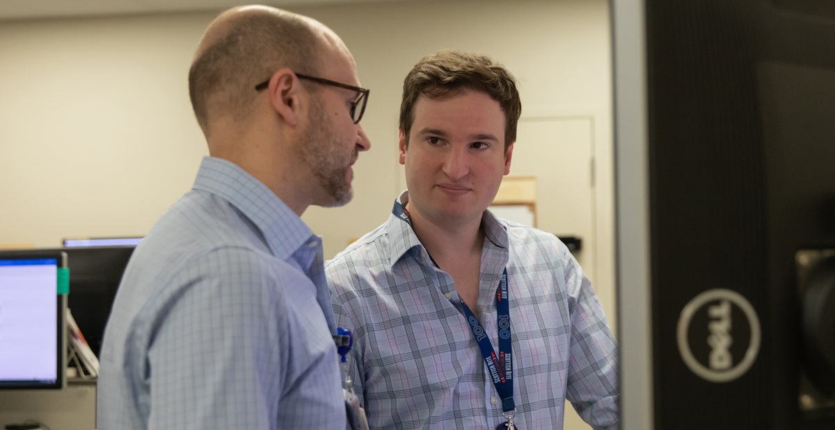 Two men in a room intently observing a computer screen, engaged in discussion or analysis of the displayed content.