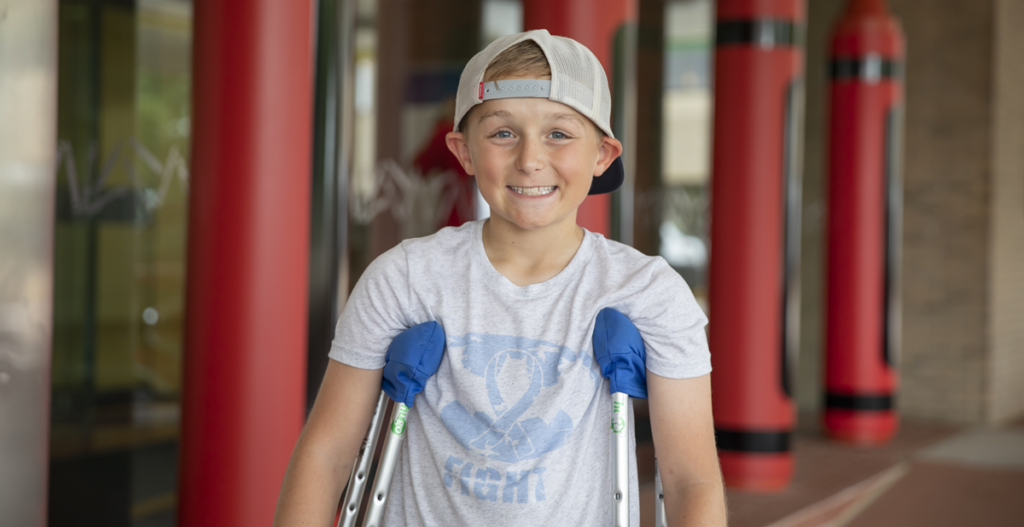 A boy in a cap smiles for the camera while standing with his crutches.