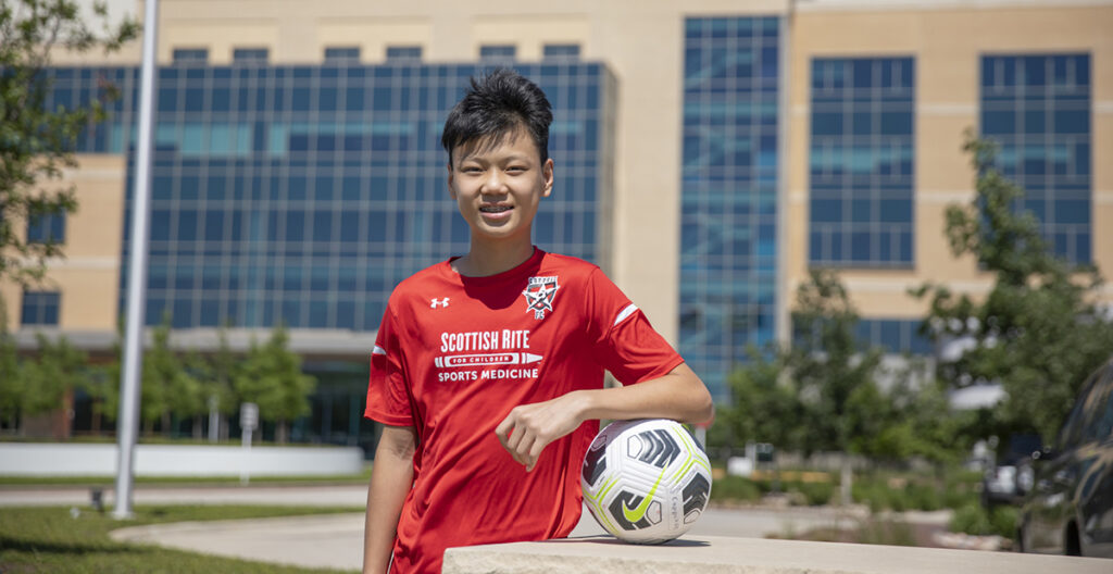 H SCOTTISH RITE נון FOR CHILDREN SPORTS MEDICINE. A boy in a scottish rite sports medicine shirt holds a soccer ball