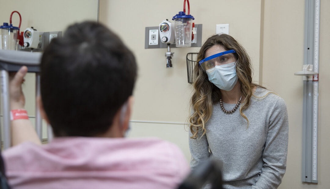 A woman wearing a face shield talks to a man in a wheelchair